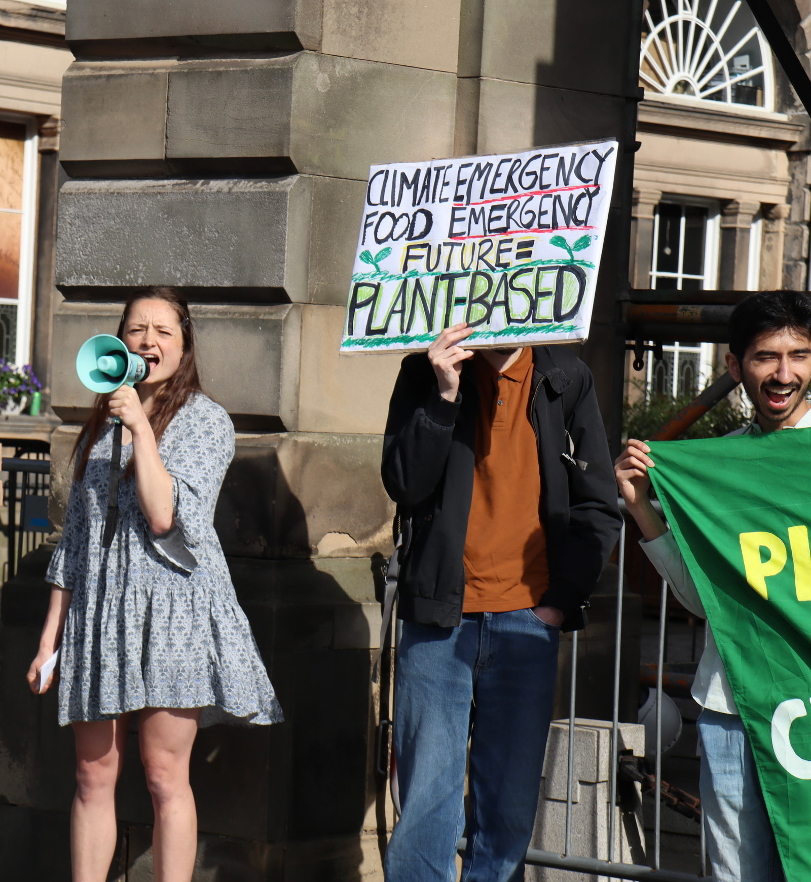 Three people stand together outdoors holding signs and a flag; one person holds a blue megaphone.