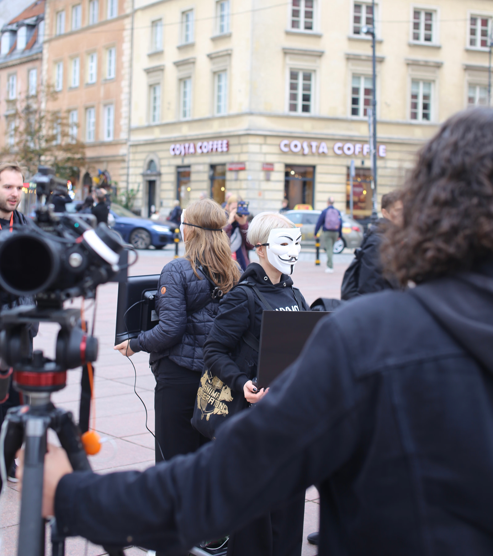 A group of people stands near a camera setup in an urban street scene with a coffee shop in the background.