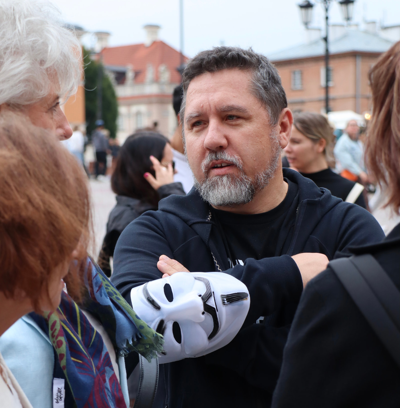 A man in a black shirt stands with arms crossed, wearing a white mask with eye holes, surrounded by several people in an outdoor public square.