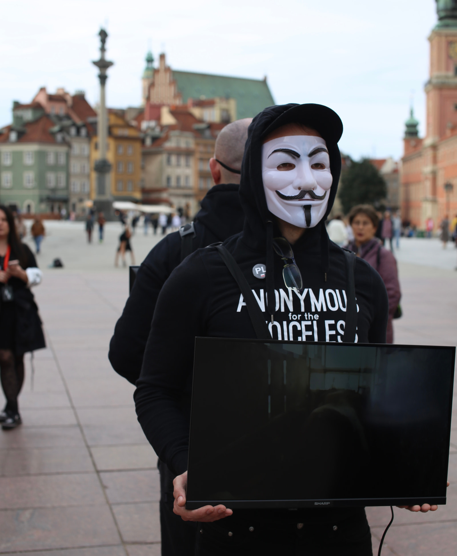 A person wearing a mask and a black hoodie holds a flat-screen monitor in a public square with historic buildings in the background.