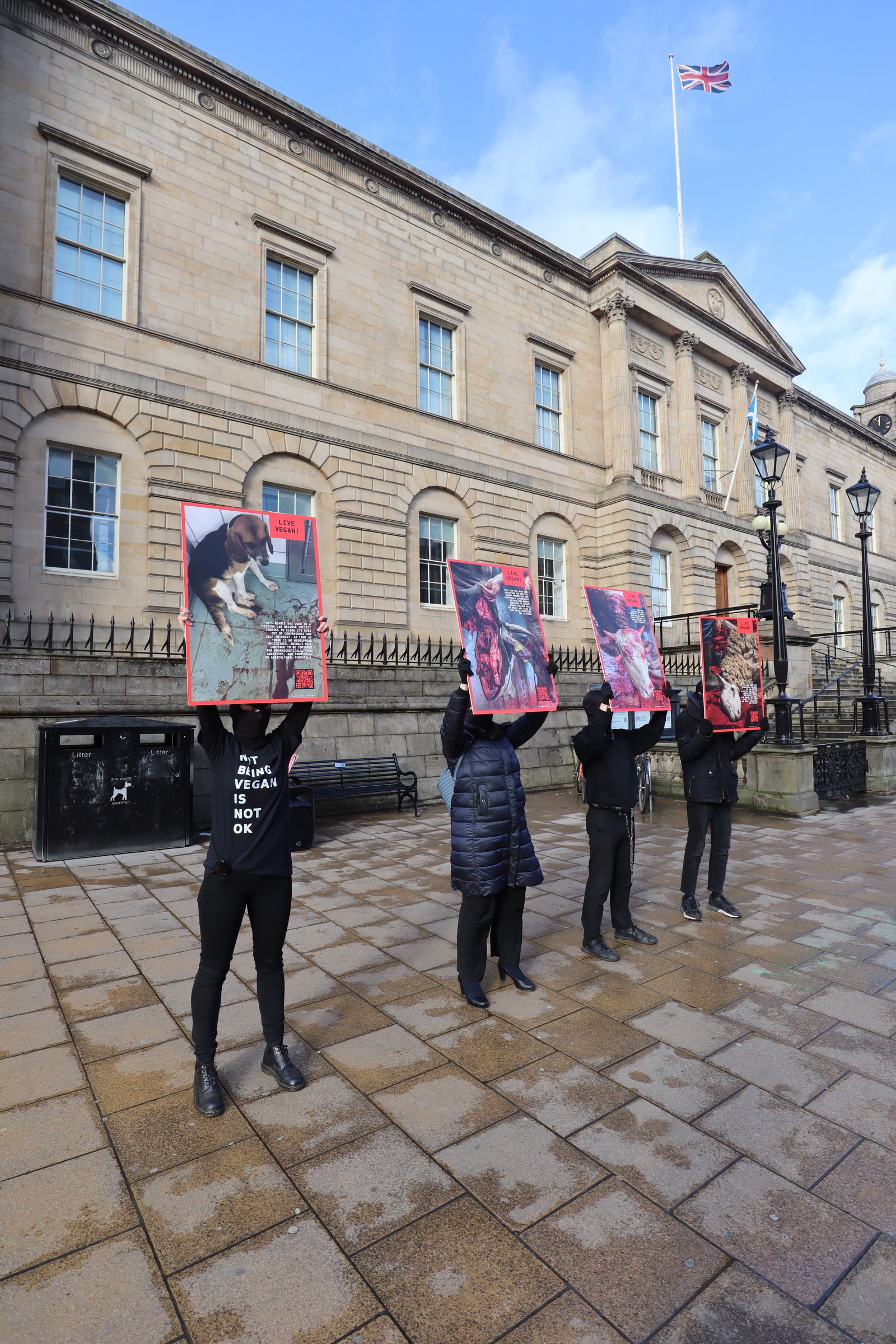 Four people stand in front of a large stone building holding graphic protest signs, with a flagpole and flag visible above them.
