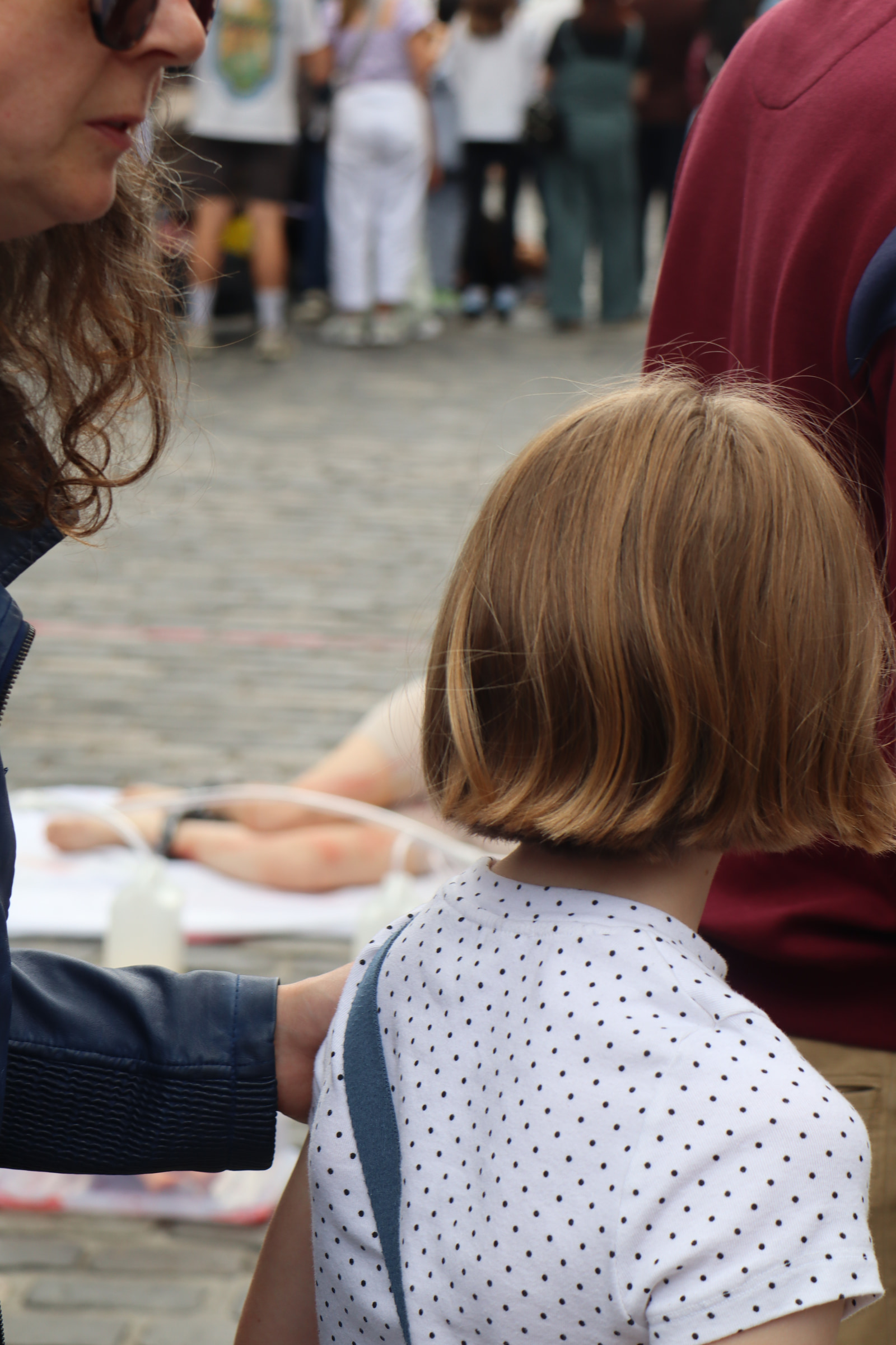A woman in sunglasses holds the arm of a child with short hair, standing in a crowded outdoor setting.