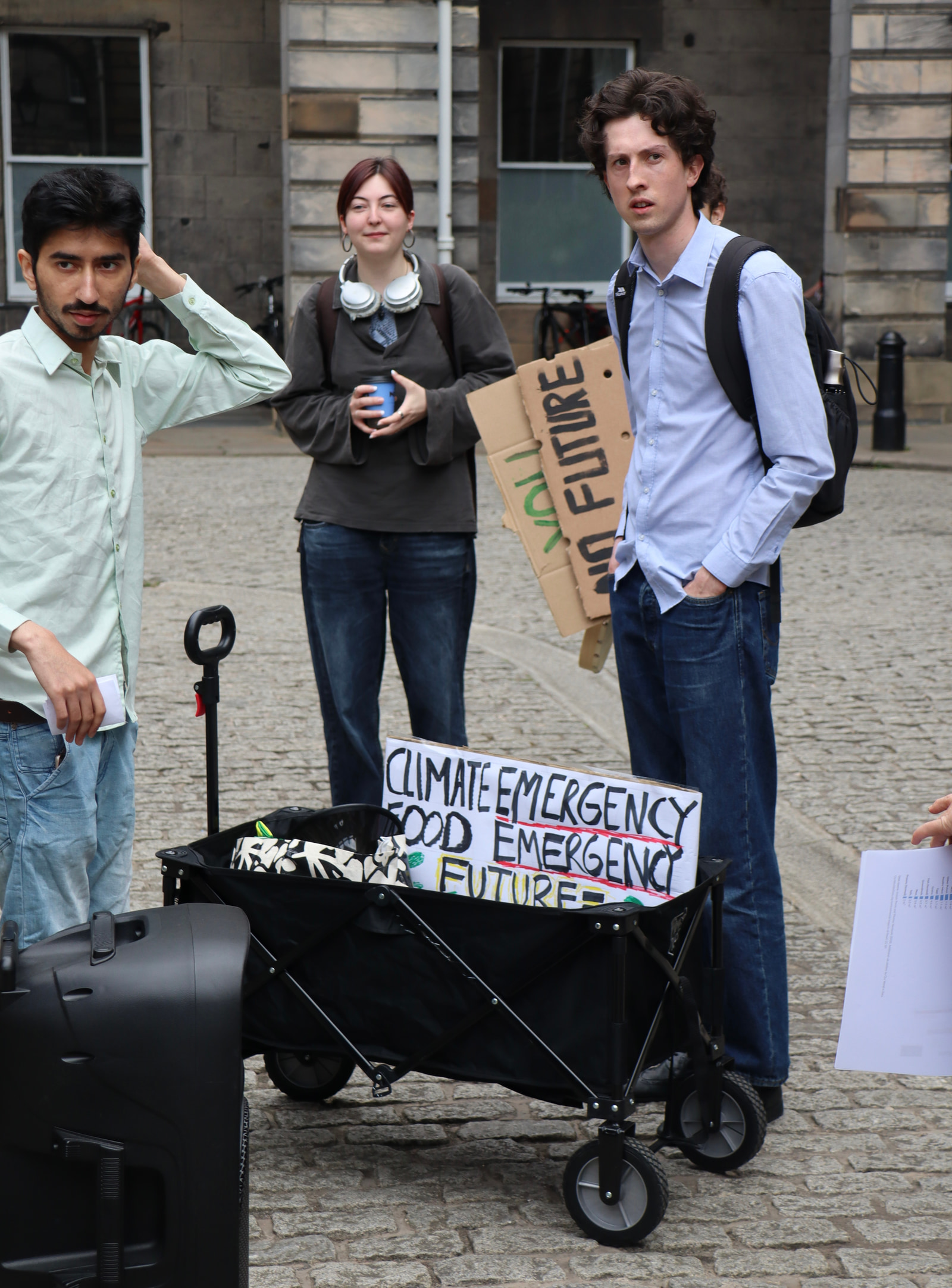 Three people stand near a wheeled cart holding signs that read "Climate Emergency Food Future" and "No Future," with a building and bicycles in the background.