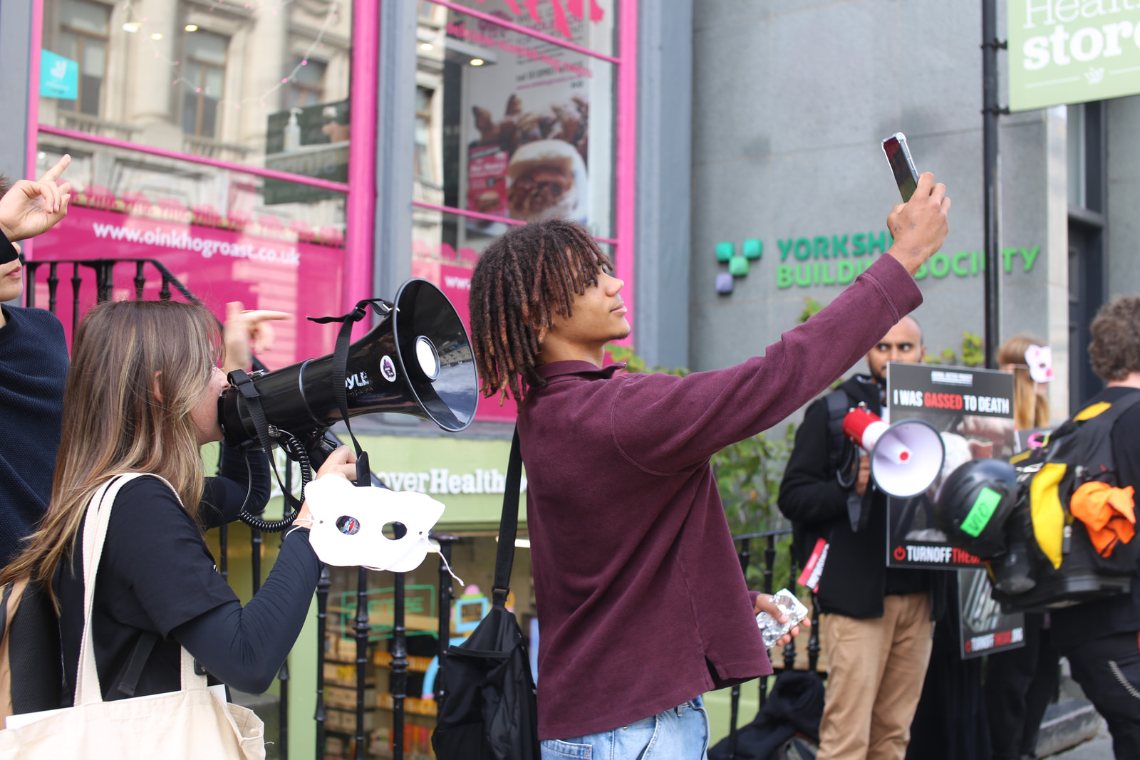 A group of people gather in a public space, some holding megaphones and signs, with one person raising a smartphone to capture the moment.