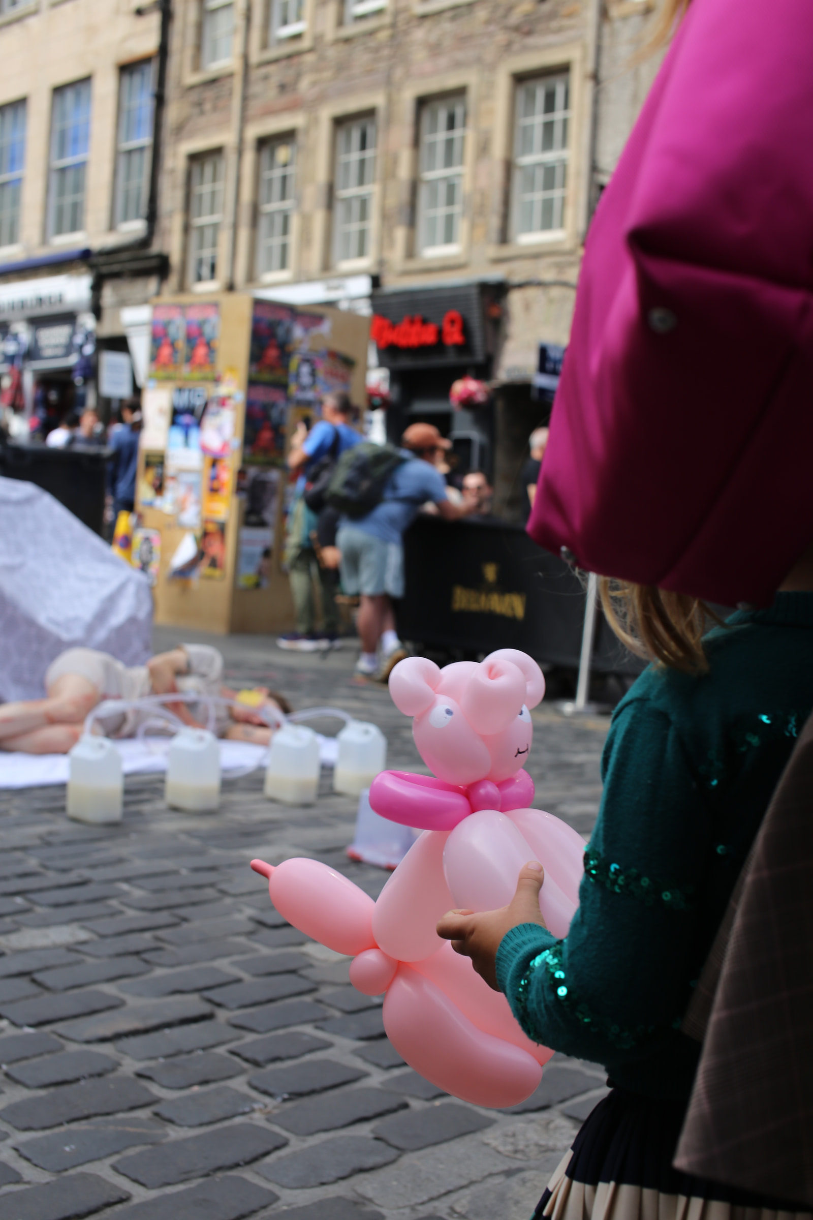 A person holds a pink balloon pig in a busy street scene with people and stalls in the background.