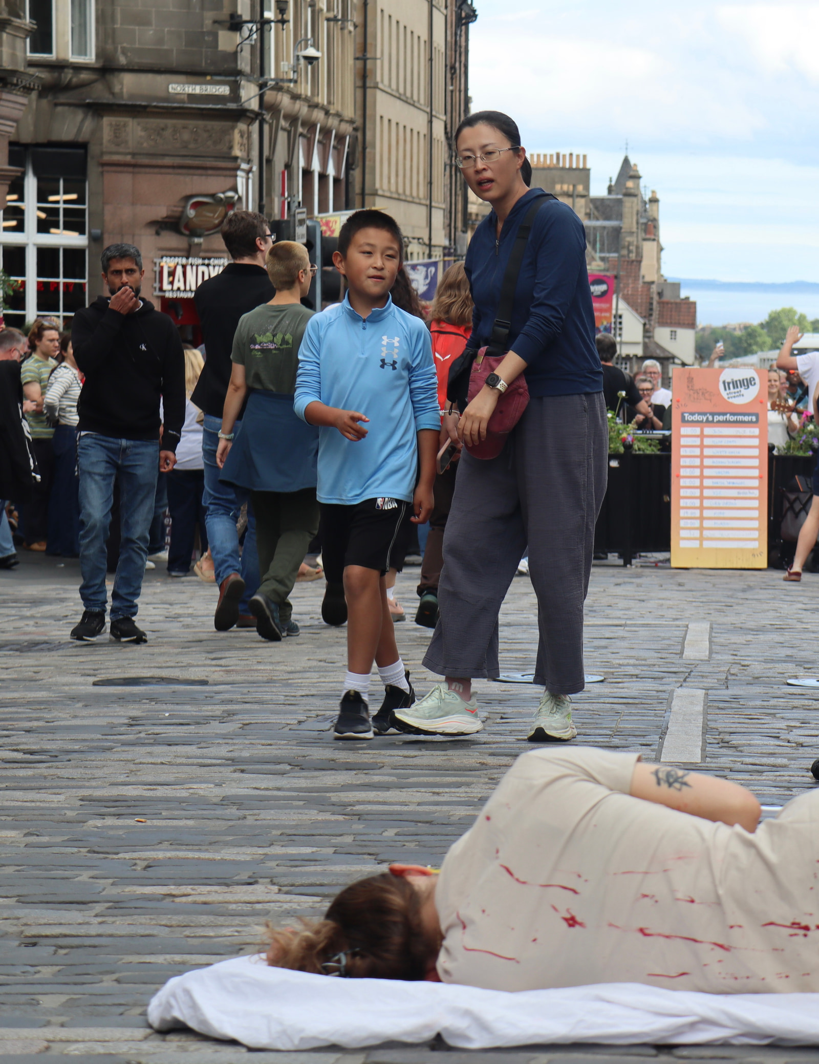 A group of people walk along a cobblestone street in an urban setting, with a person lying on the ground covered in a white cloth and red stains nearby.