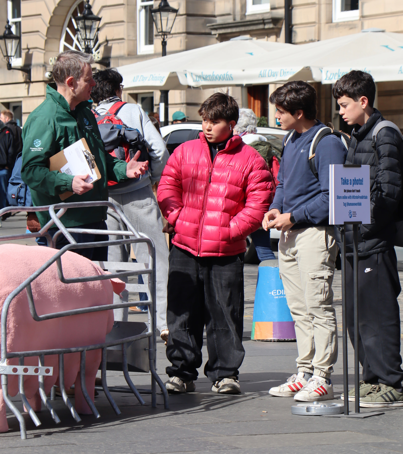 Three young people stand together on a city street near a sign that says "Take a photo!" while an older man in a green jacket talks to them.