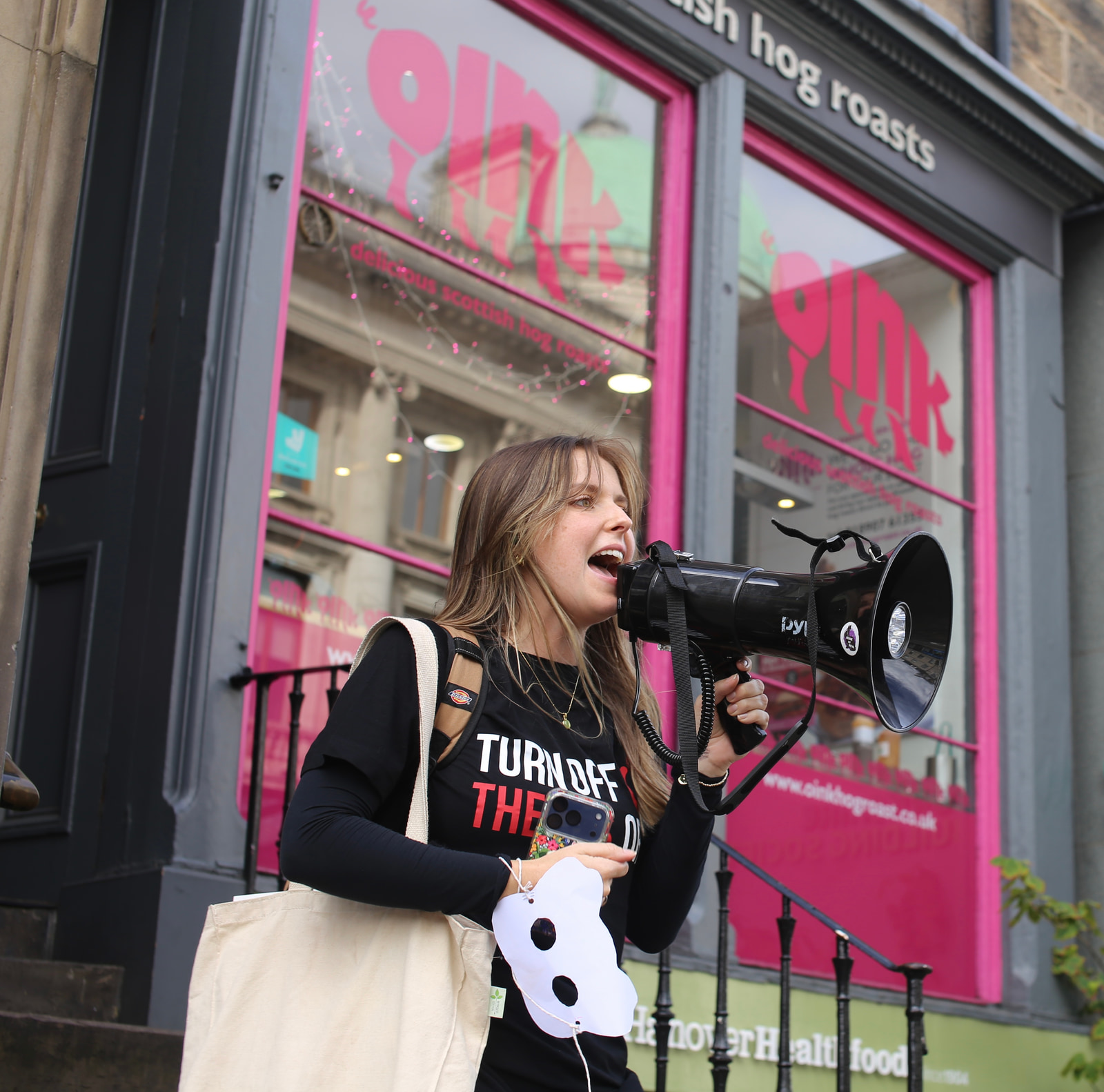 A person holds a megaphone and speaks, wearing a black long-sleeve shirt with a printed slogan, in front of a storefront with bright pink signage.