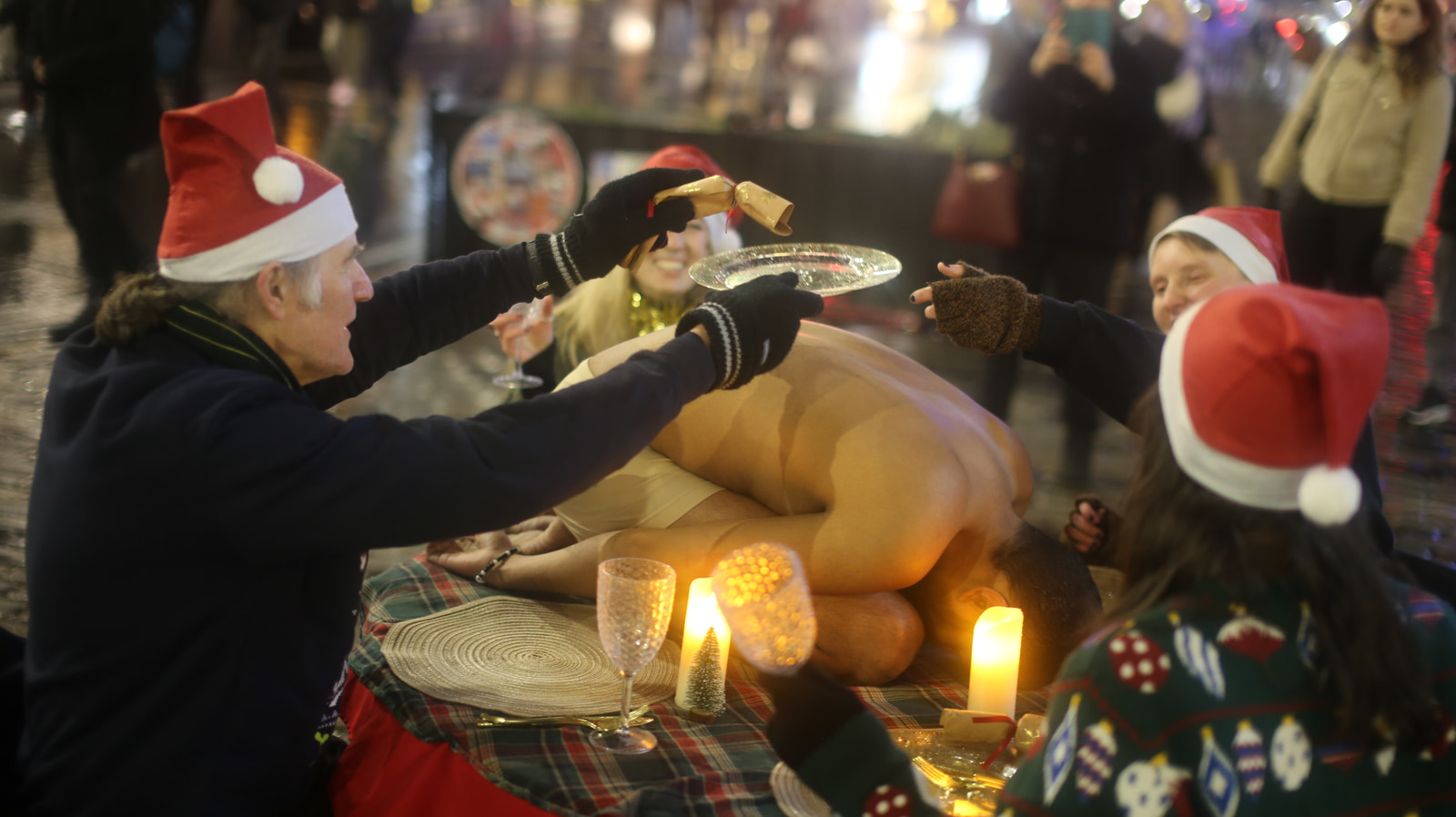 People in festive hats gather around a large inflatable reindeer, holding glasses and a plate, with candles and a plaid blanket on the ground.