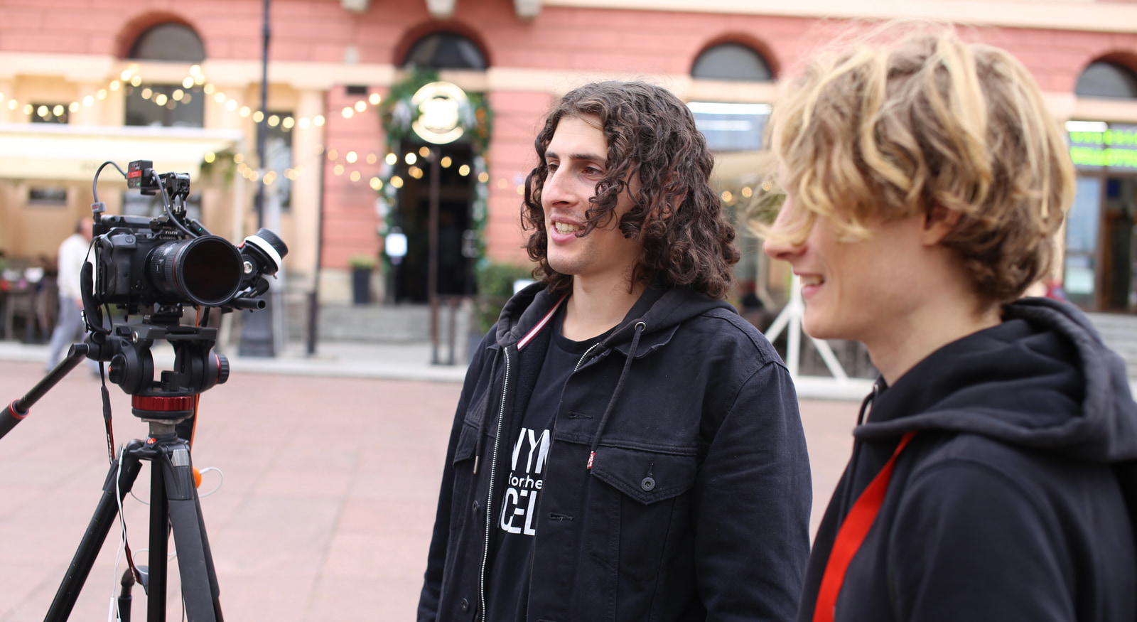 Two people stand together, one holding a camera on a tripod, in an urban square with festive lights and a decorated building behind them.