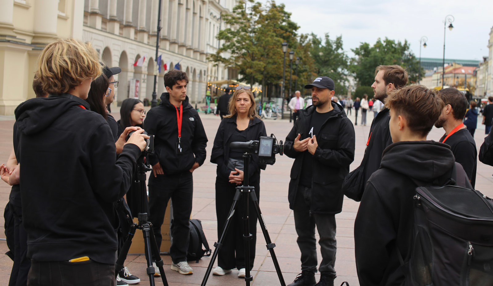 A group of people stands together in an open public square, with one person speaking to the others while a camera on a tripod is positioned in front of them.