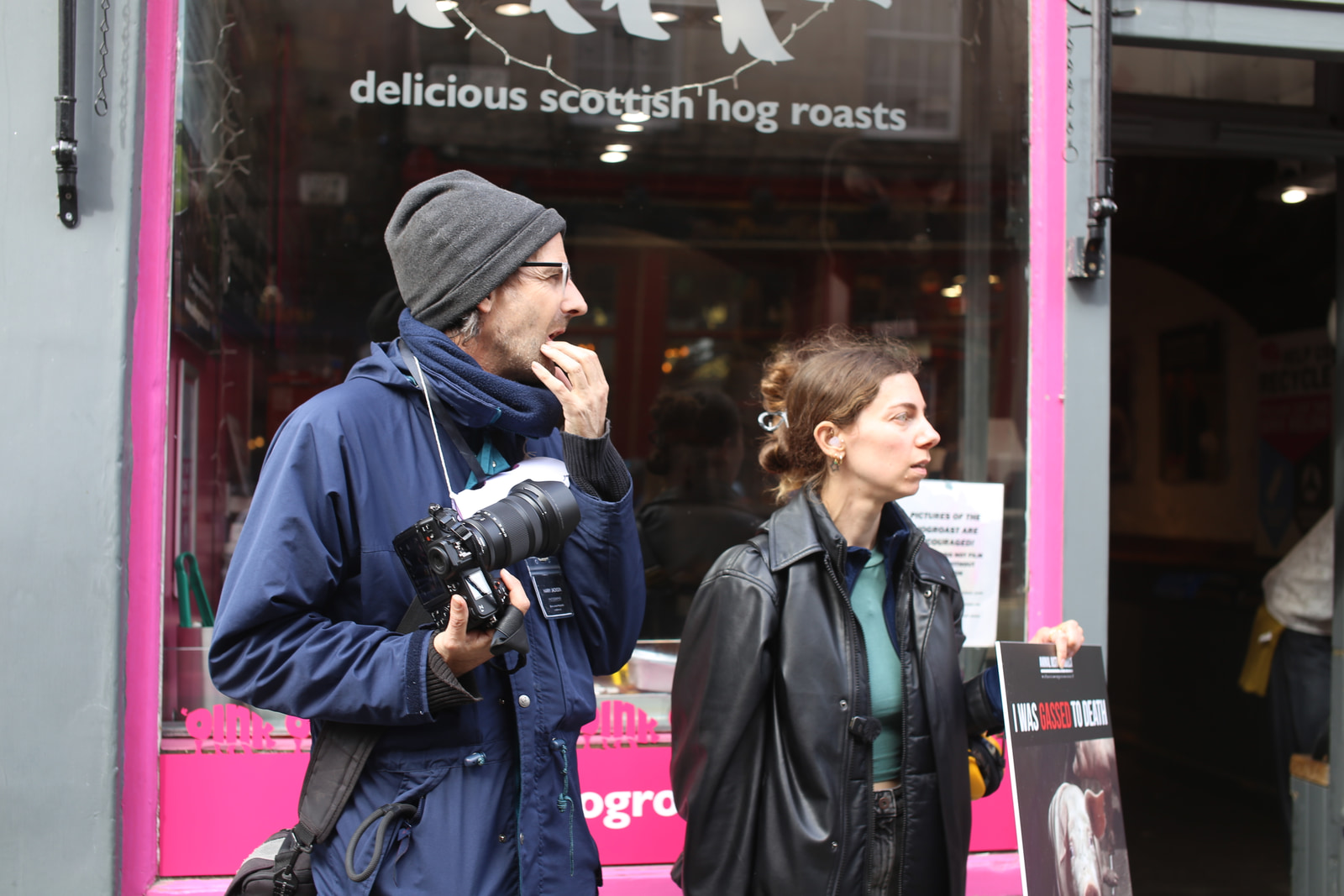 A man and a woman stand outside a storefront, the man holding a camera and the woman holding a printed sign.