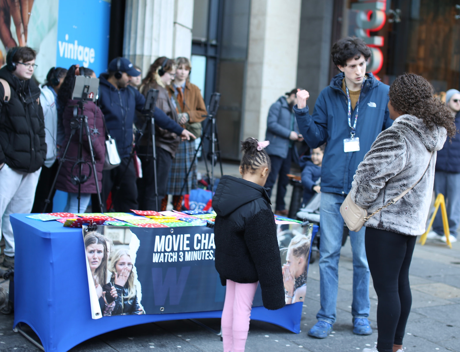 A group of people stands near a promotional table with a poster, while a child in pink pants looks at the display.