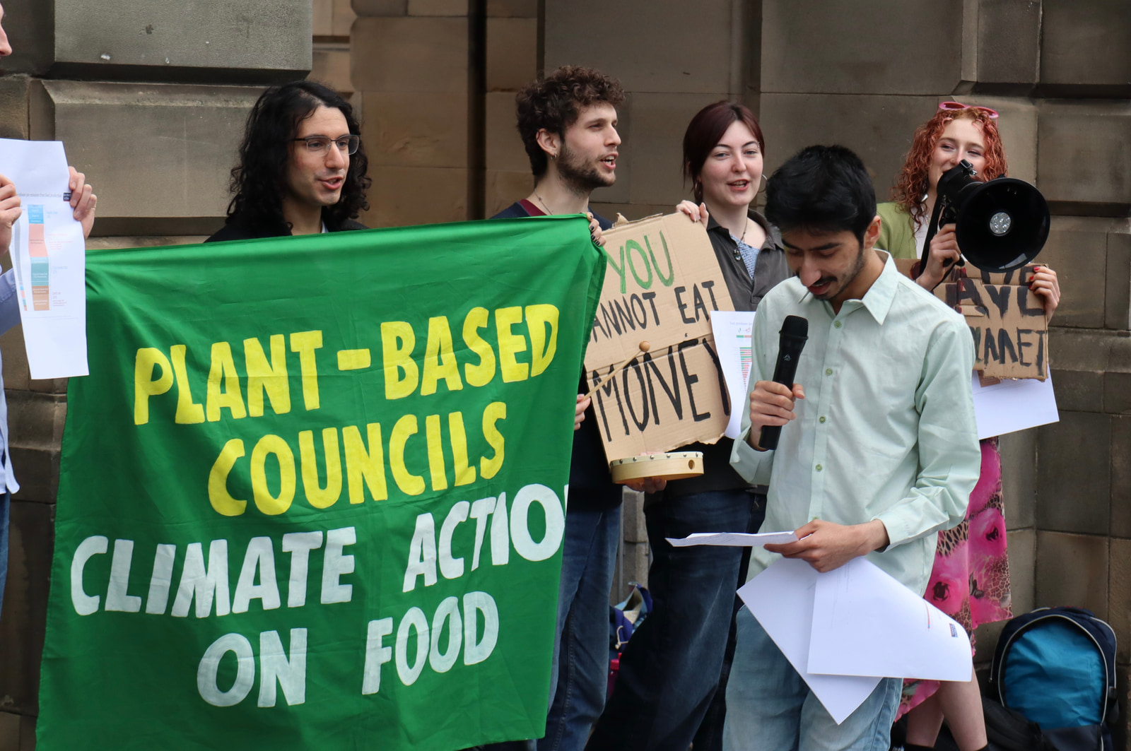 A group of people stand together holding signs and a banner advocating for plant-based food policies, with one person speaking into a microphone.