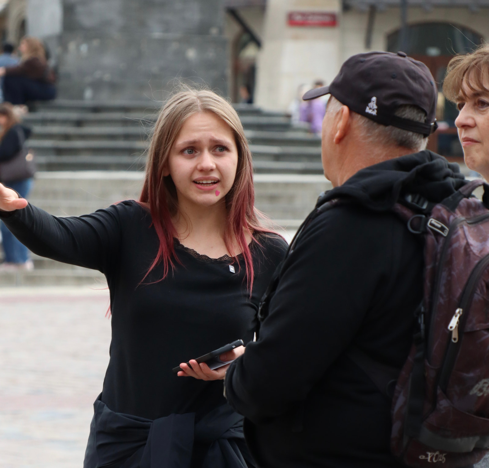 A young woman with pink highlights in her hair gestures toward the camera while standing next to an older man and woman on a set of stone steps.